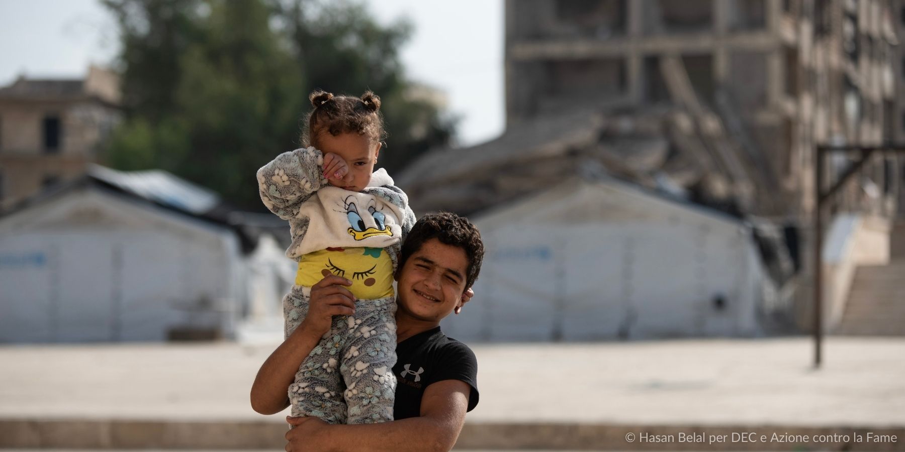 Terremoto in Siria e Turchia del 2023, un bambino tiene in braccio la sorellina con sfondo di macerie
